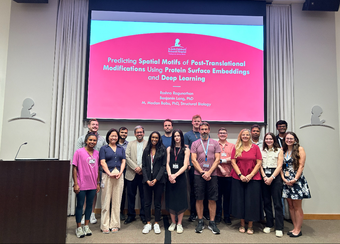 Elias standing on a stage with other researchers standing alongside her. Behind the group of researchers is a screen displaying a presentation that reads "Predicting Spatial Motifs of Post-Translational Modifications Using Protein Surface Embeddings and Deep Learning