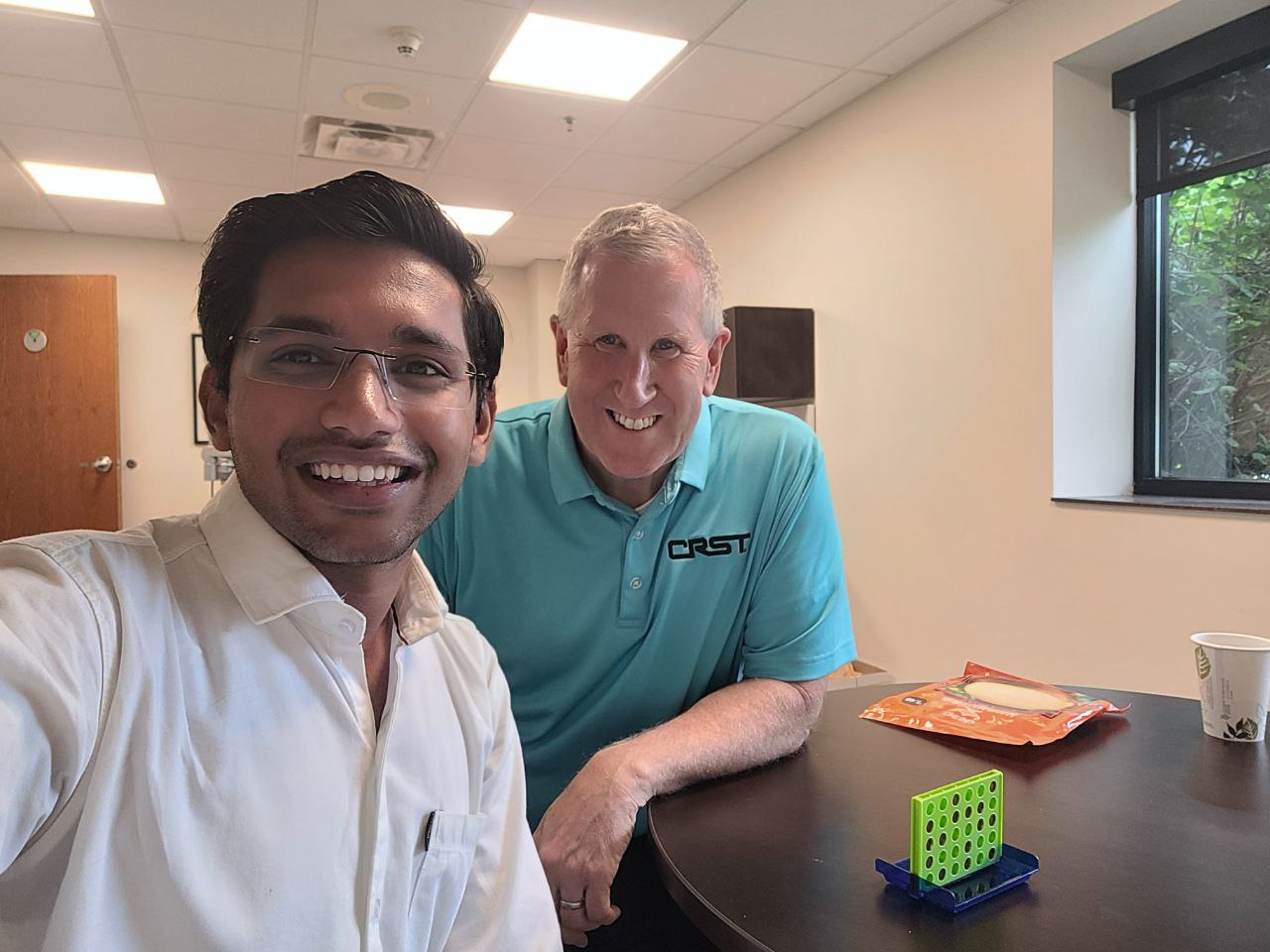 Nahar takes a selfie with the new CEO of CRST, Mike Gannon. A mini connect-4 game is on a table in front of the pair. 