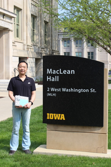 Hantao Zhang pictured holding wrapped retirement present from his peers in front of MacLean Hall sign on Pentacrest between MLH and Shaeffer Hall.