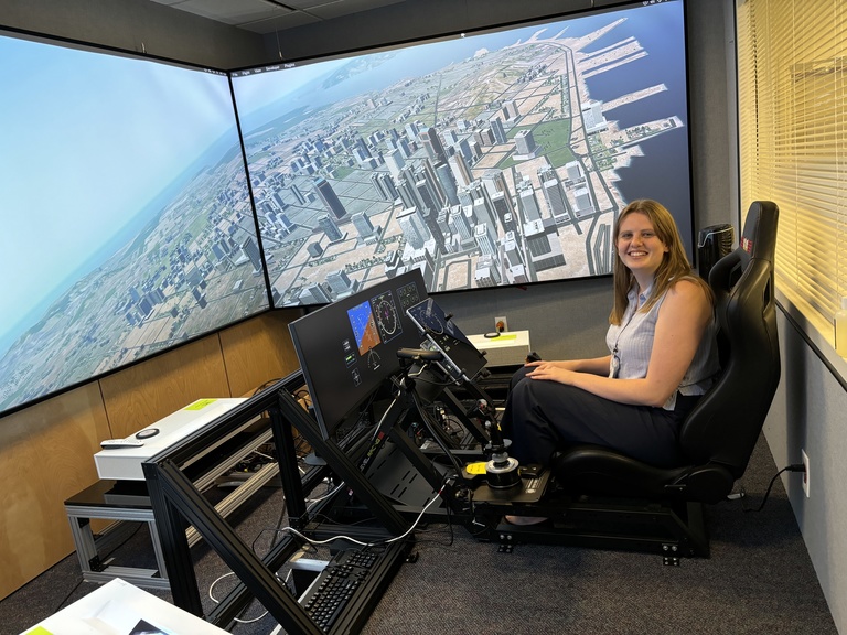 Paulsen sitting in front of two large screens displaying an overhead view of a simulated city and two smaller screens mimicking the dashboard of an aircraft. 