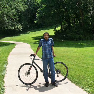Photo of Abdal Mohamed on a path surrounded by green lawn, standing in front of a grey bicycle.