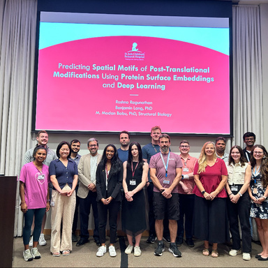 Elias standing on a stage with other researchers standing alongside her. Behind the group of researchers is a screen displaying a presentation that reads "Predicting Spatial Motifs of Post-Translational Modifications Using Protein Surface Embeddings and Deep Learning