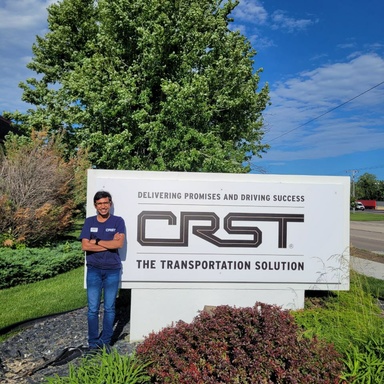 Veetrag Nahar standing outside in front of a sign reading "Delivering Promises and Driving Success: CRST - The Transportation Solution"