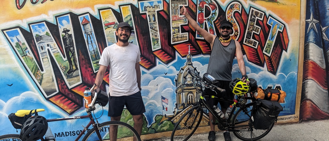 Hussam and Osama celebrating reaching Winterset - in from of "Welcome to Winterset" postcard-style mural, after an 80 miles day