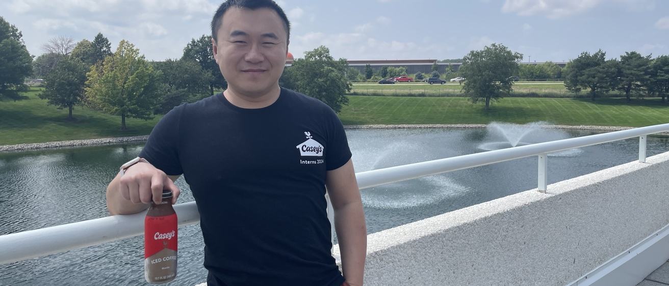 Cai standing on a bridge near Casey's corporate headquarters, wearing a Casey's shirt and holding a Casey's iced coffee