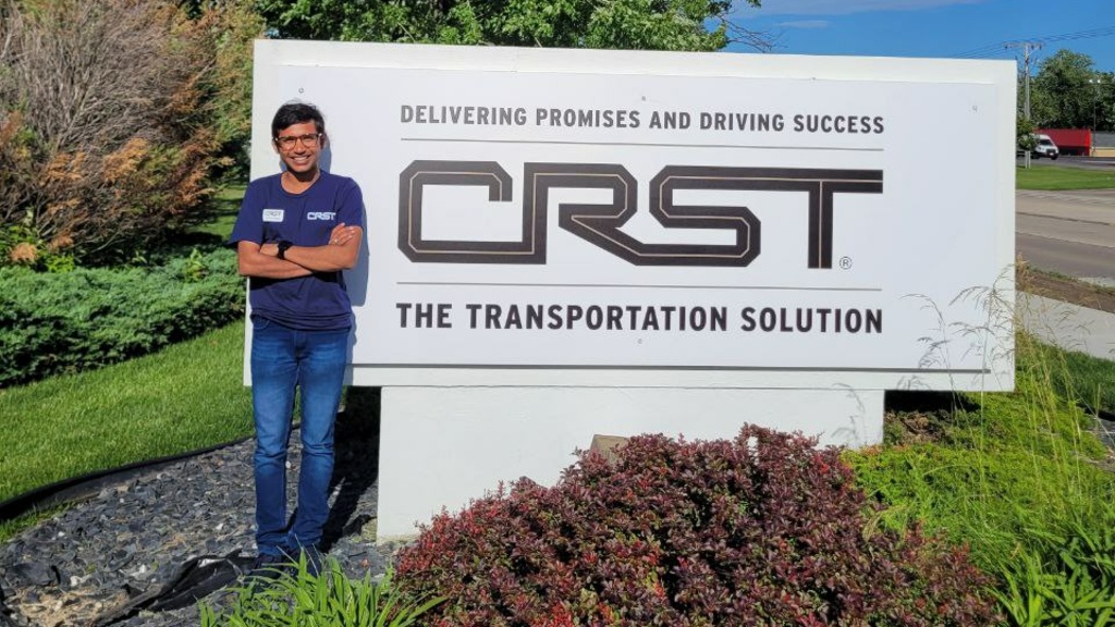 Nahar standing next to a sign outside reading "Delivering Promises and Driving Success: CRST - The Transportation Solution"